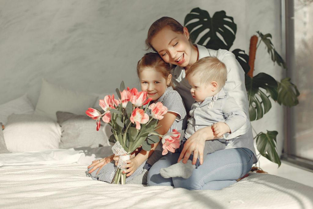 Cheerful mother with daughter and son sitting on bed and playing speaking with baby while spending time together at home