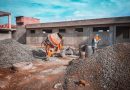 A construction site with a worker wearing safety gear next to a cement mixer and gravel piles.