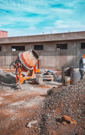A construction site with a worker wearing safety gear next to a cement mixer and gravel piles.