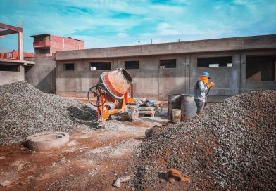 A construction site with a worker wearing safety gear next to a cement mixer and gravel piles.