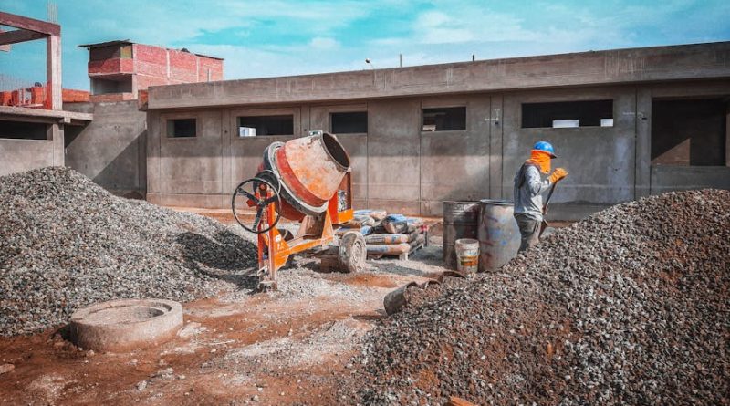 A construction site with a worker wearing safety gear next to a cement mixer and gravel piles.