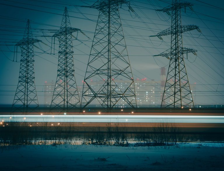 Moody night photo of electric towers silhouetted against an urban cityscape, highlighting powerful infrastructure.