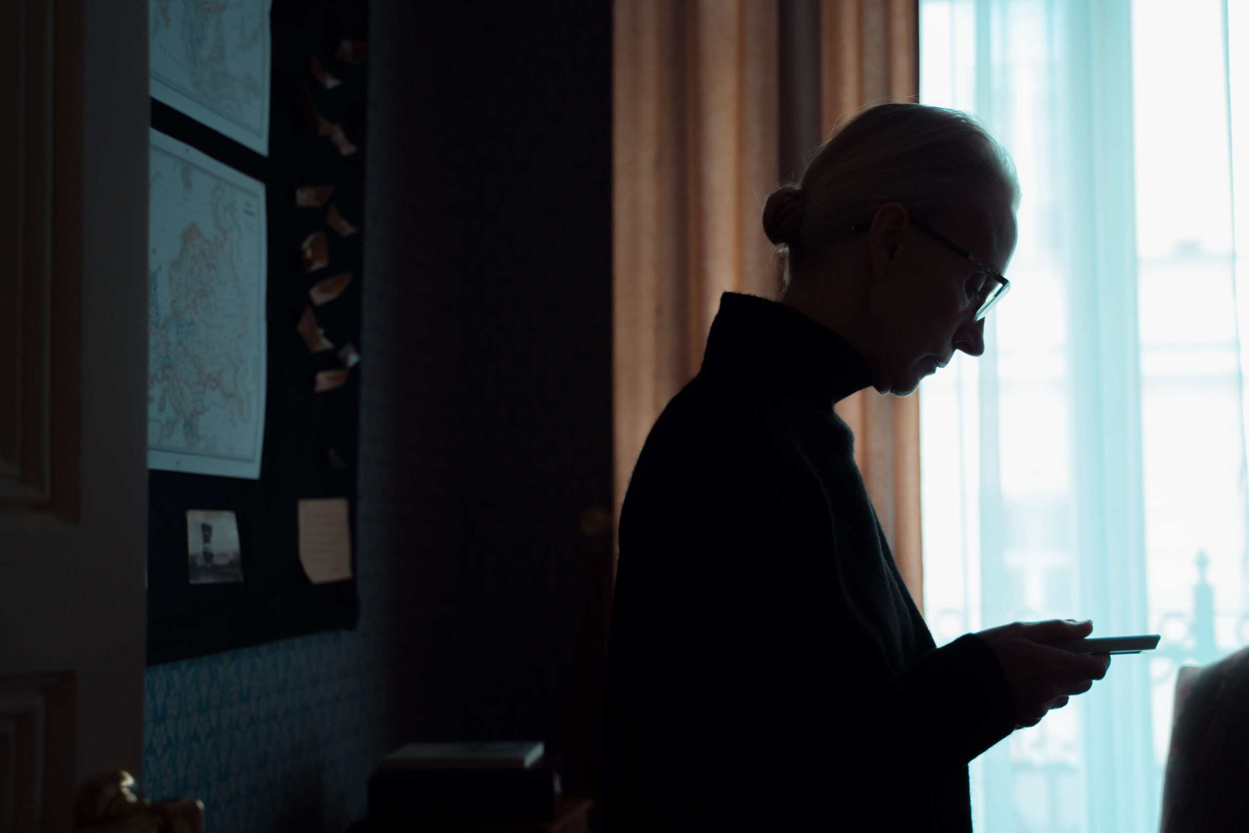 Silhouette of a senior woman looking at a smartphone in a dimly lit room, conveying introspection and solitude.